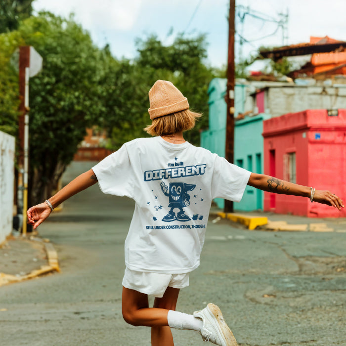Person wearing a white t-shirt with text and graphics, standing on a street with colorful buildings in the background.