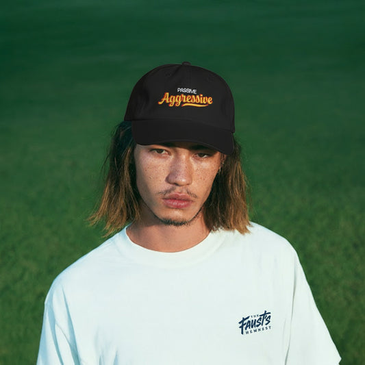Person wearing a black cap with 'Aggressive' text and a light blue shirt with 'Trusts' text, standing against a green background.