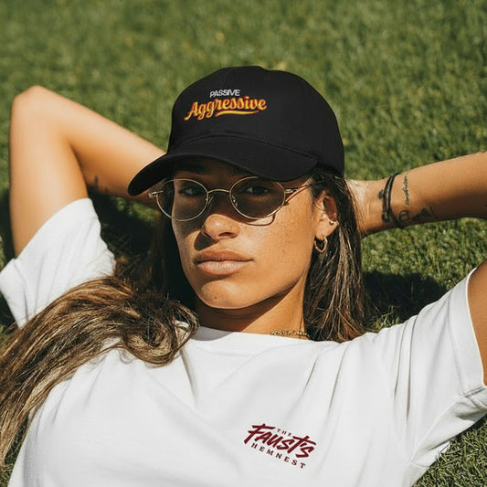 Person wearing a black cap with 'Passive Aggressive' text and a white t-shirt with 'Faust's' logo, lying on grass.