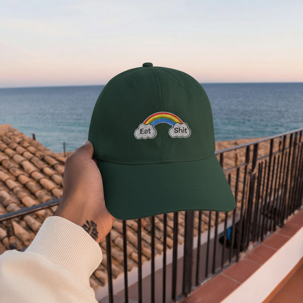 Green cap with rainbow and cloud design held by a hand against a scenic ocean backdrop