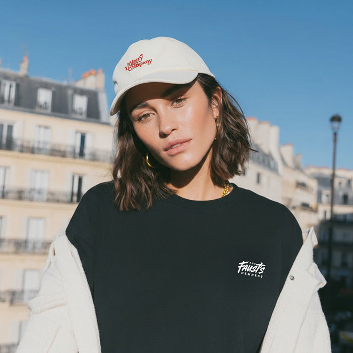 Woman wearing a black t-shirt with a logo and a white cap with red text, standing in an urban setting.
