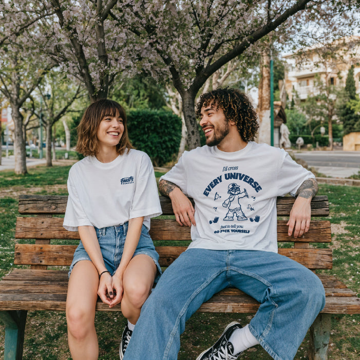 Two people sitting on a bench in a park, wearing white t-shirts with graphic designs.