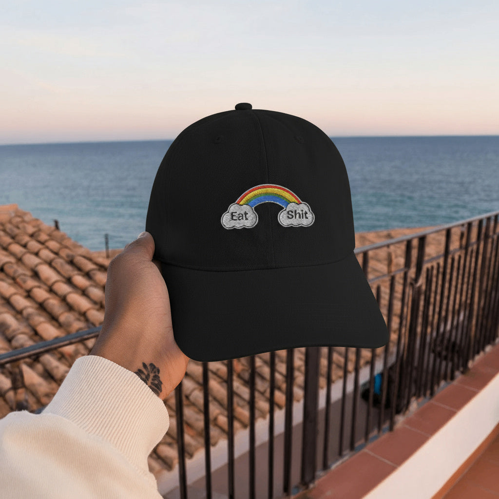 Black cap with rainbow and text held by a hand against a scenic ocean backdrop