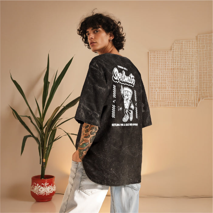 Person wearing a black t-shirt with graphic design, standing in a room with a plant and wall art.