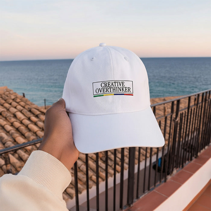 White cap with 'Creative Overthinker' text held by a hand against a scenic ocean backdrop