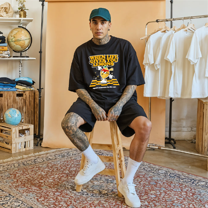 Person sitting on a stool wearing a black t-shirt with a graphic design, black shorts, white socks, and white shoes in a room with a clothing rack and shelves.
